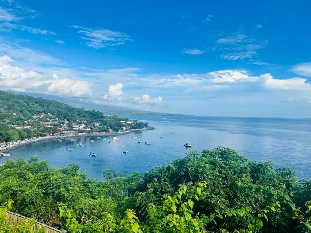 Panoramablick auf die Jemeluk Bay in Amed, Bali, mit türkisblauem Wasser, Fischerbooten (Jukung) und der grünen Küstenlinie unter blauem Himmel.